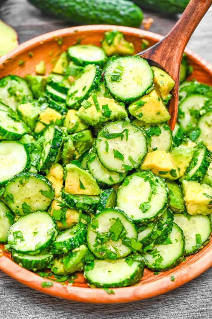Cucumber avocado salad in a bowl with fresh herbs and a wooden spoon