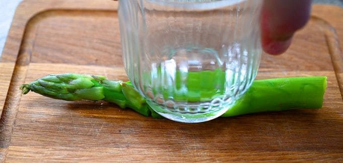 Smashing a blanched asparagus spear with the bottom of a glass on a wooden cutting board.