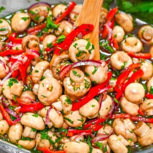 Zesty Garlic Marinated Mushrooms with sliced red bell peppers, purple onions, and fresh herbs being stirred in a glass bowl with a wooden spoon.