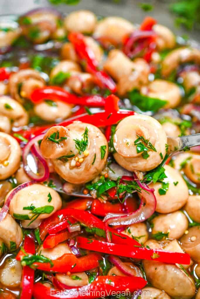 Zesty Garlic Marinated Mushrooms with sliced red bell peppers, purple onions, and fresh herbs, shown in a close-up where a metal spoon lifts mushrooms from the glossy marinade.