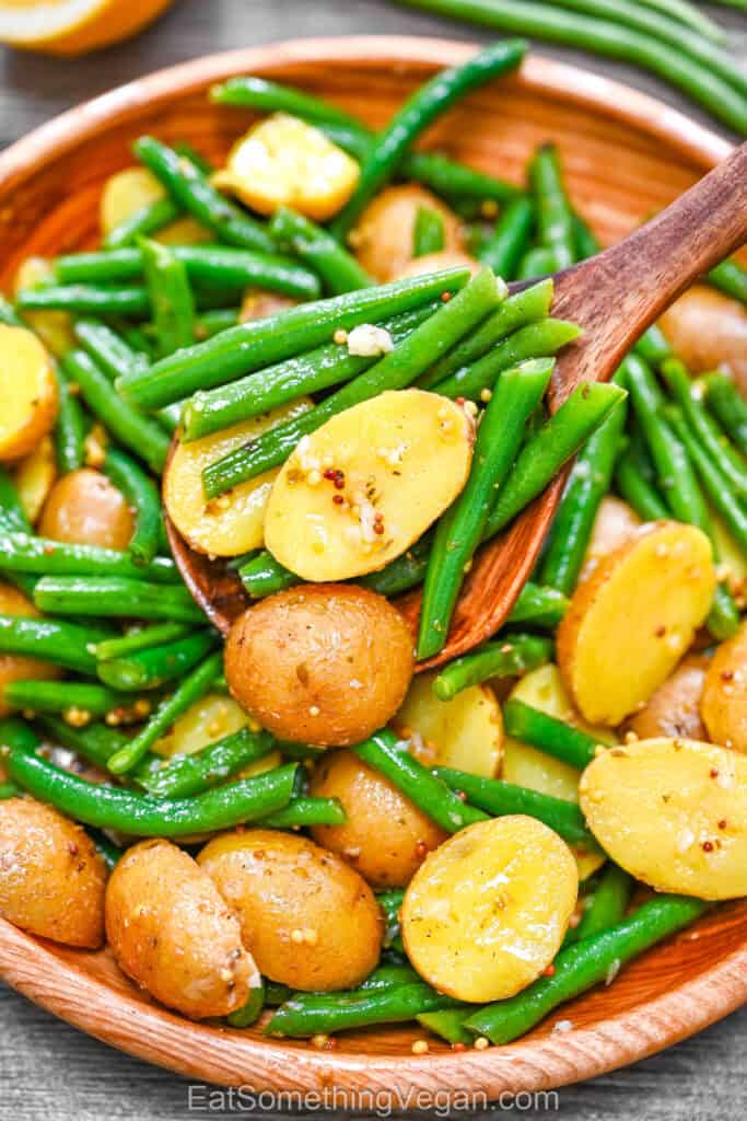 Warm Potato and Green Bean Salad served in a wooden bowl, being tossed with a wooden spoon to mix the tender potatoes and crisp green beans.