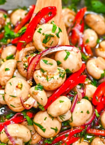 Close-up of marinated whole button mushrooms with bright red bell peppers, purple onions, and fresh herbs in a zesty garlic dressing.