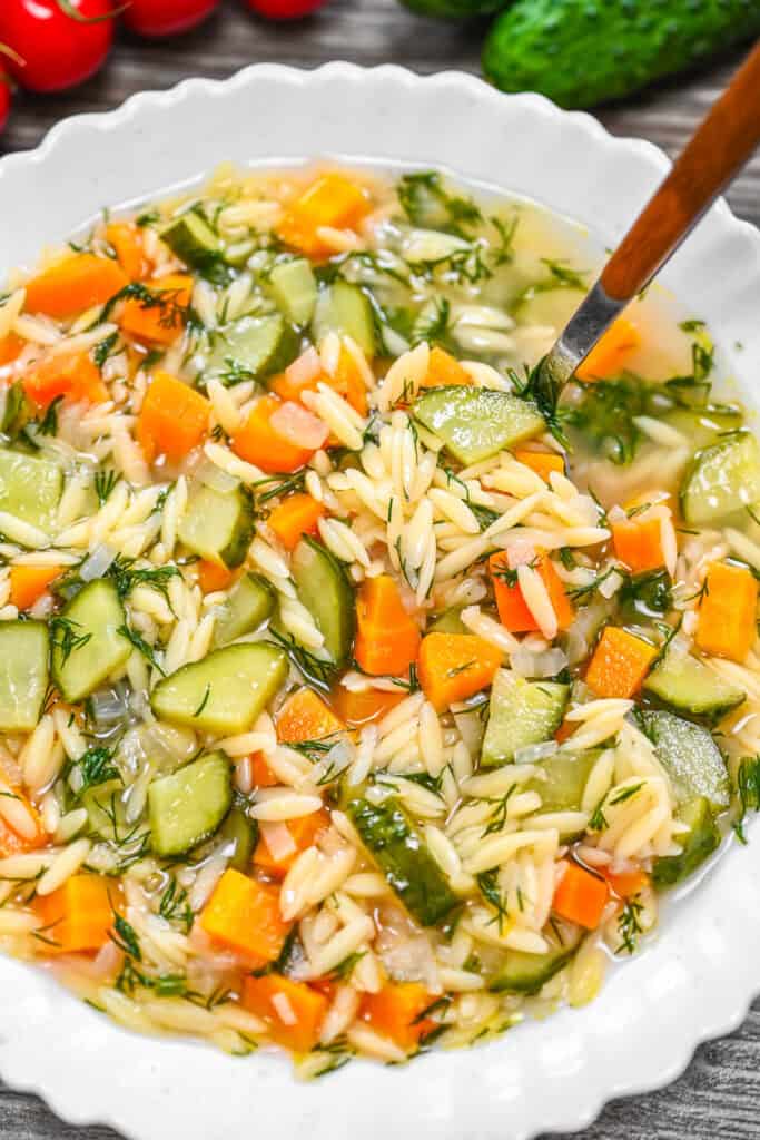 Cucumber Orzo Soup served in a scalloped white bowl with a wooden-handled spoon.