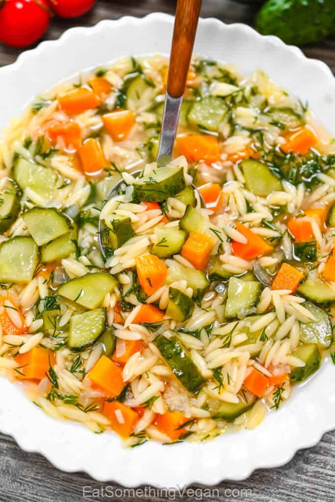 Cucumber Orzo Soup in a scalloped white bowl with a wooden-handled spoon stirring the soup.