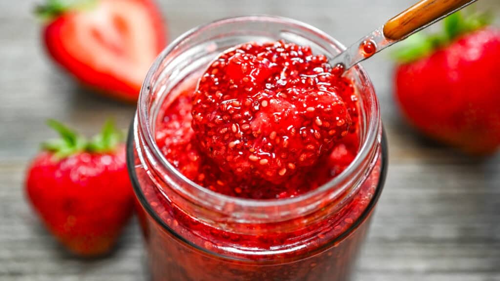 3-Ingredient Strawberry Chia Jam in a jar close-up, showing a spoon lifting the thick, seed-filled jam on a wooden surface.