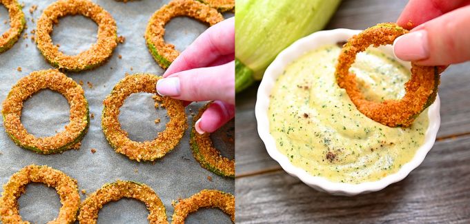 A hand picking up a baked crispy zucchini ring from a baking sheet and dipping it into a small white bowl filled with creamy green sauce.
