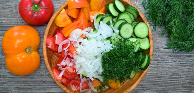 A wooden bowl filled with chopped red and yellow tomatoes, sliced cucumbers, onions, and fresh dill, with whole tomatoes on the side.