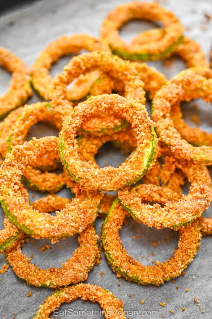 Crispy zucchini rings piled on a baking sheet lined with parchment paper, showing a golden-brown breaded texture.