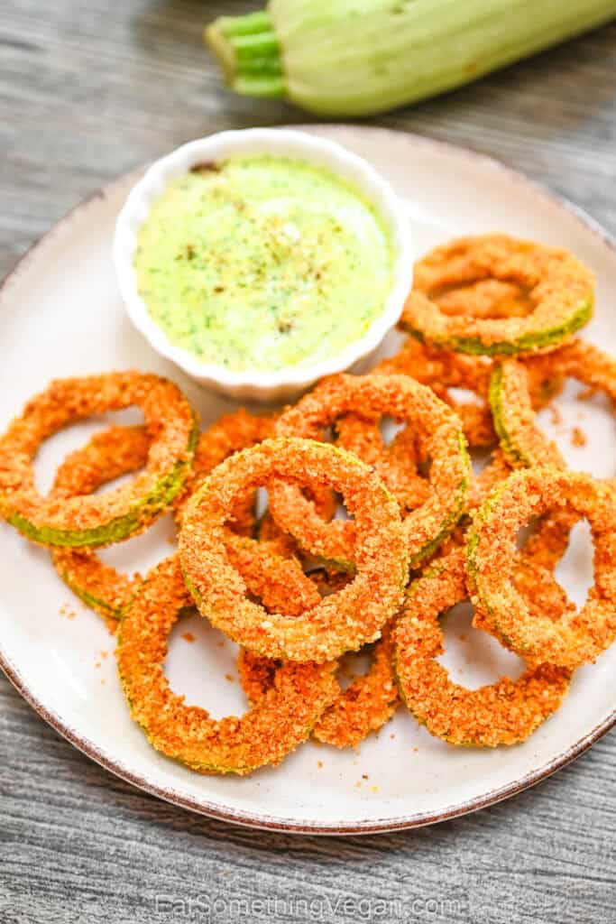 Crispy zucchini rings served on a white plate with a small bowl of creamy green dipping sauce.