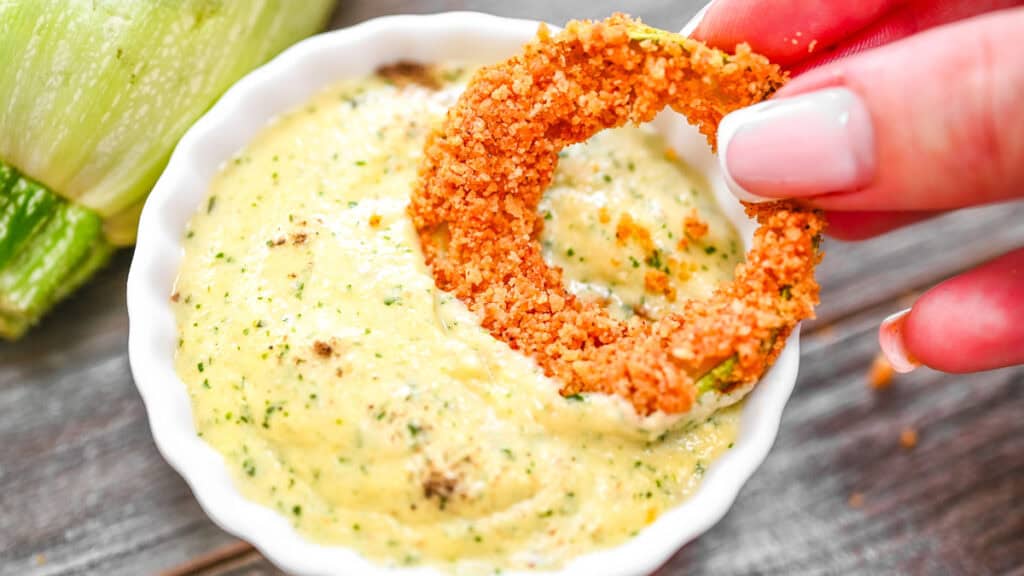 Crispy zucchini ring being dipped into a small white bowl of creamy green herb sauce by a hand.