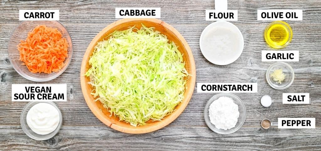 Top-down view of ingredients for making vegan cabbage fritters, including shredded cabbage, grated carrot, flour, cornstarch, vegan sour cream, garlic, olive oil, salt, and pepper.