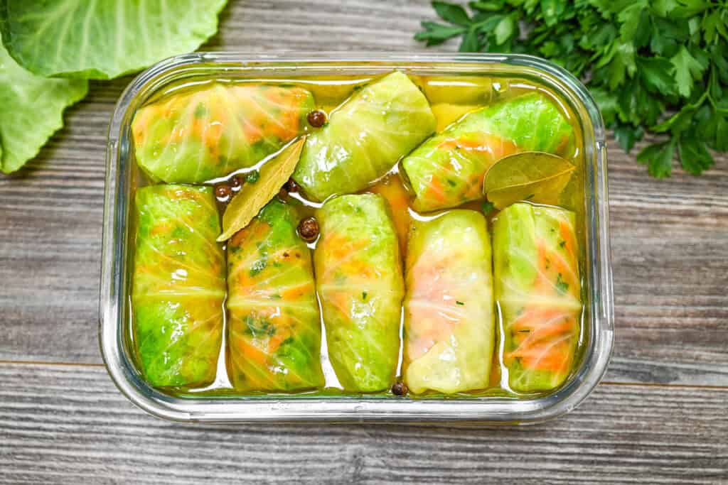 A top-down view of marinated pickled cabbage rolls in a sealed glass container with visible bay leaves and brine on a rustic wooden table.