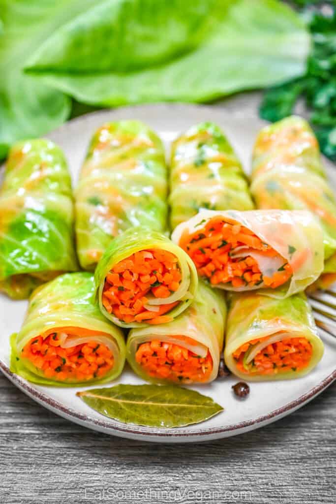 A plate of pickled cabbage rolls, with several rolls cut open to reveal a vibrant shredded carrot and herb filling, served on a rustic wooden table with a bay leaf.
