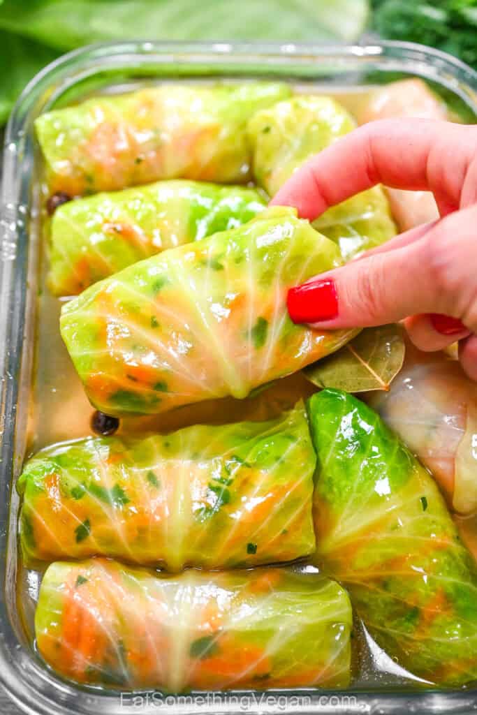 A hand taking a marinated pickled cabbage roll from a glass container filled with brine and bay leaves
