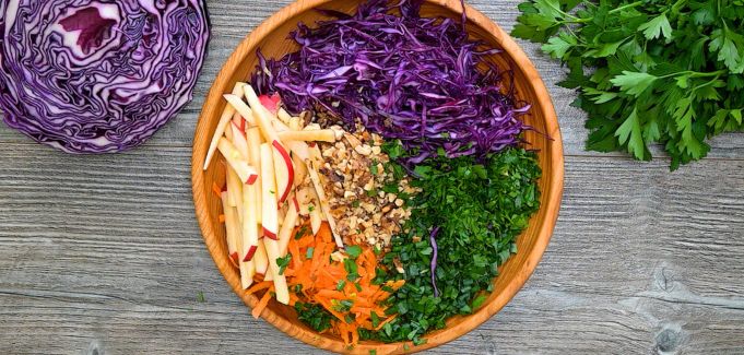 Shredded red cabbage, sliced apples, carrots, walnuts, and herbs in a wooden mixing bowl.