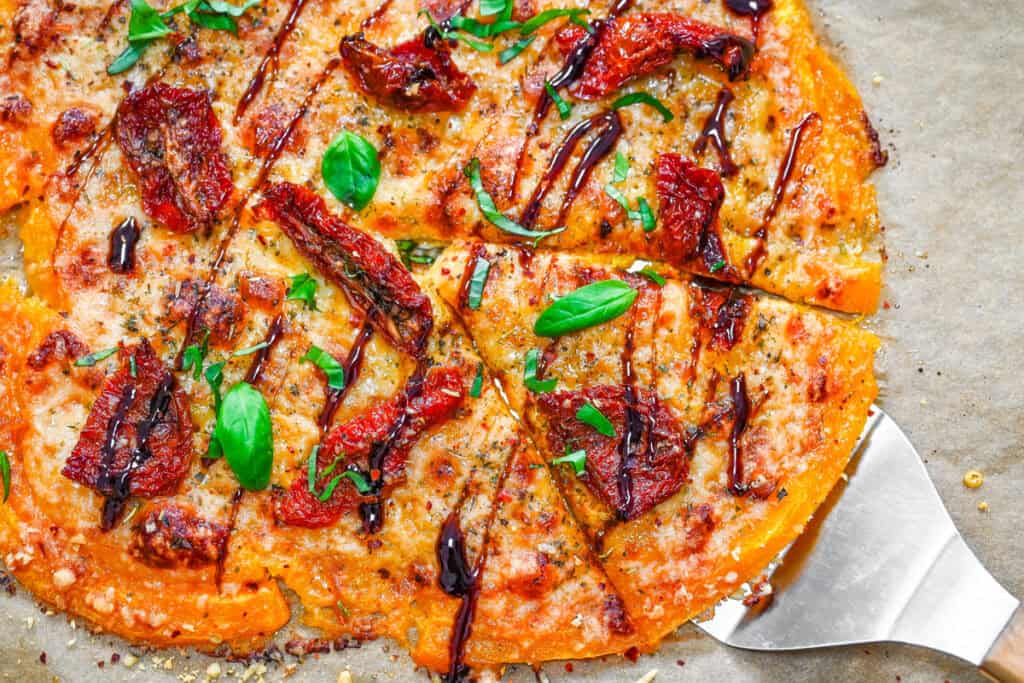 Smashed Butternut Squash Pizza crust cut into portions with a spatula sliding under a golden, herb-sprinkled slice.