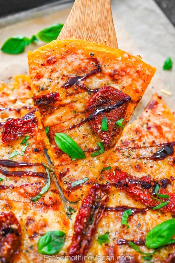 Smashed Butternut Squash Pizza slice lifted with a wooden spatula, topped with melted cheese, sun-dried tomatoes, fresh basil, and balsamic glaze.