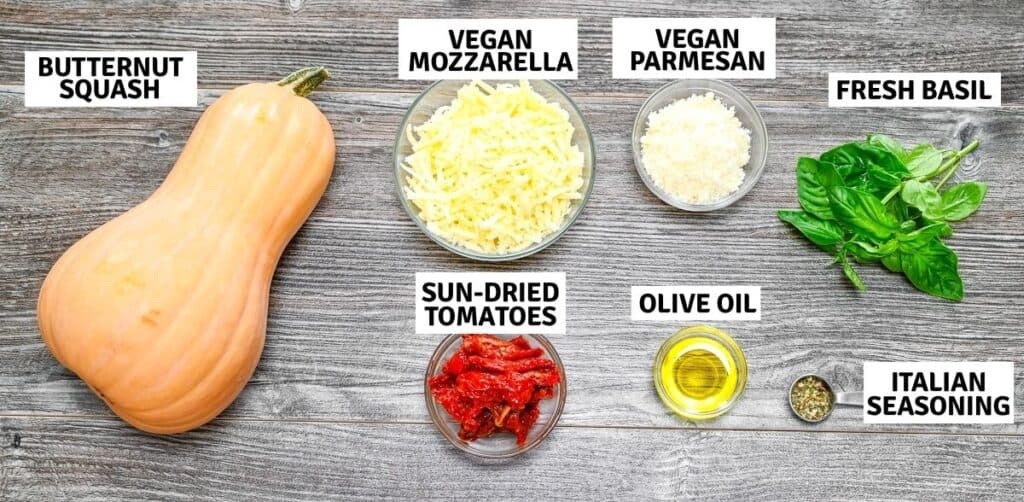 Ingredients laid out on a wooden surface including whole butternut squash, shredded dairy-free mozzarella, parmesan, fresh basil, sun-dried tomatoes, olive oil, and Italian seasoning.