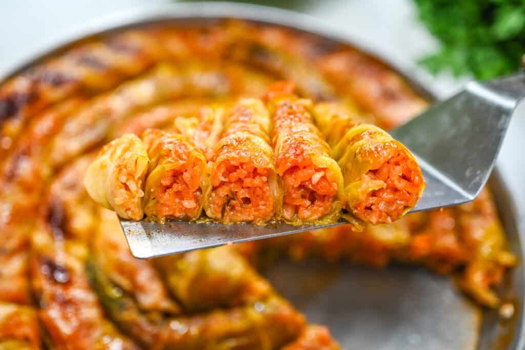 A close-up cross section of baked spiral cabbage rolls lifted on a spatula, showing the seasoned tomato rice filling inside tender cabbage layers.