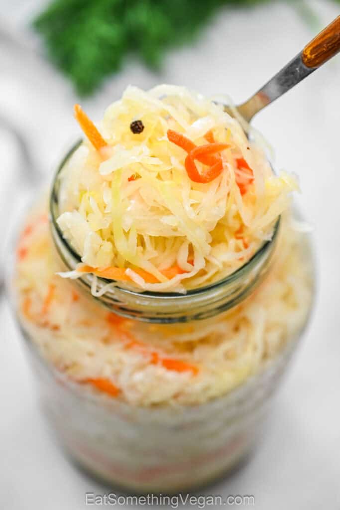 Close-up of crispy homemade sauerkraut on a fork lifted from a glass jar with shredded carrots and black peppercorn.