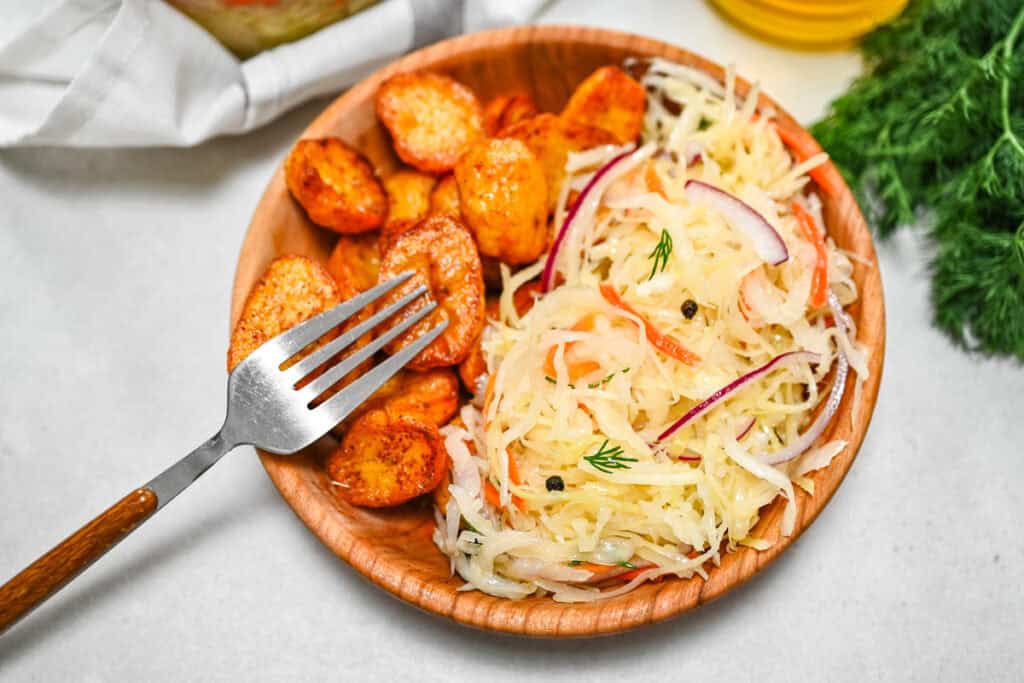Homemade crunchy sauerkraut salad with red onion and fresh dill served alongside crispy roasted potatoes in a wooden bowl with a fork.